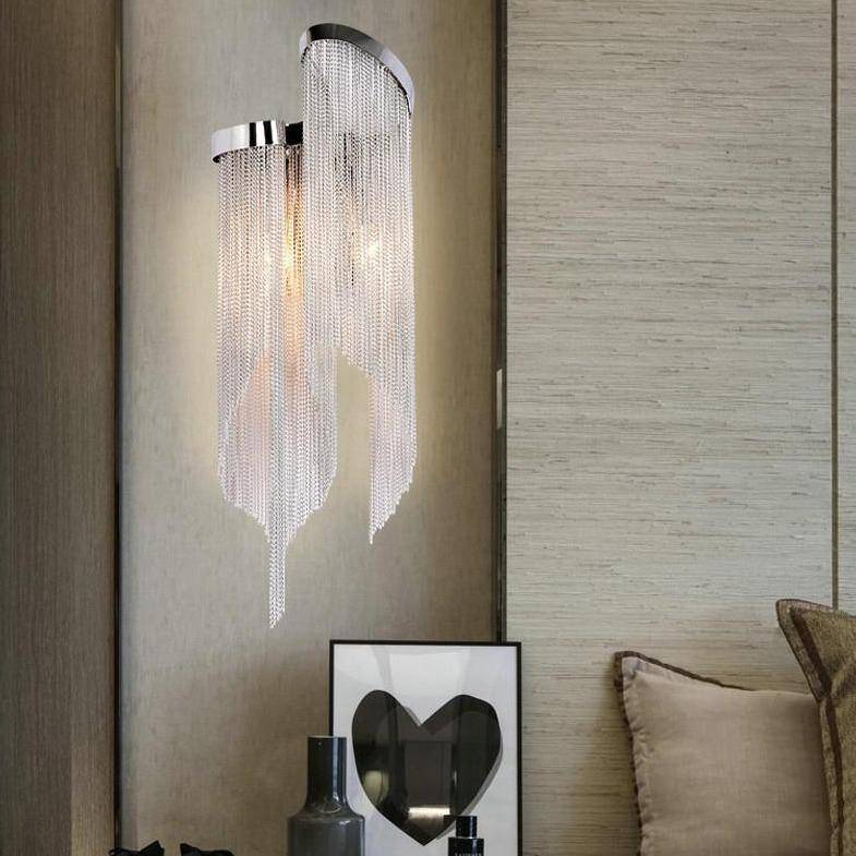 The Cadena chain tassel sconce from Morsale.com mounted beside a bed, 23-inch metal tassel drape glowing warm.