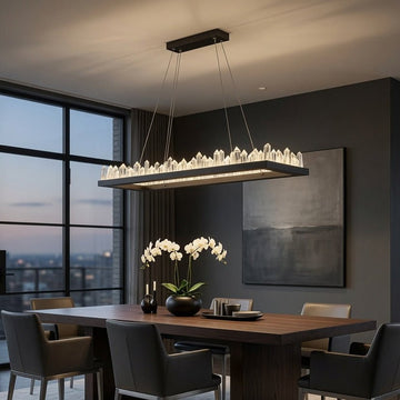 A modern rectangular dining room with a dark wood table, gray upholstered chairs, a large black chandelier with clear crystal-like prisms, and large floor-to-ceiling windows overlooking a city skyline at dusk.